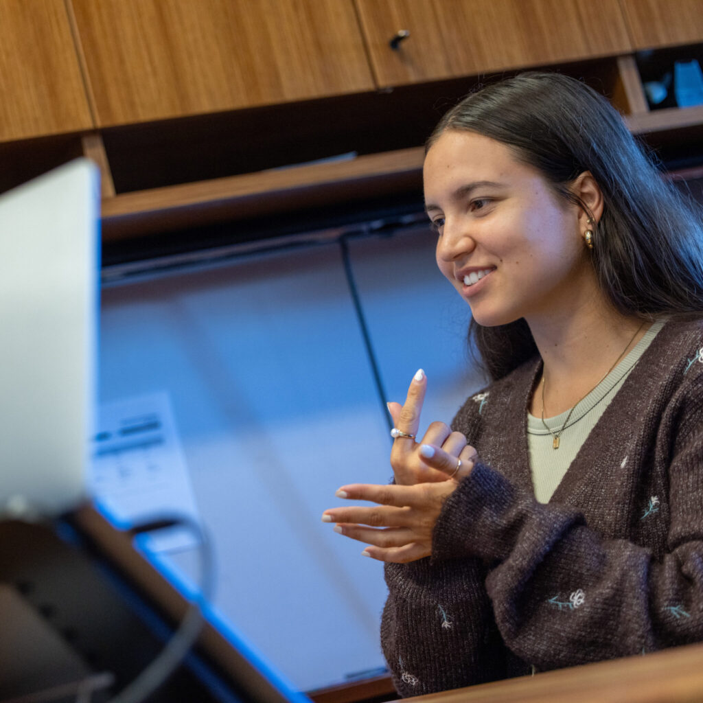 A person sits in front of a laptop, smiling and signing "graduate". They are in a well-lit office setting.