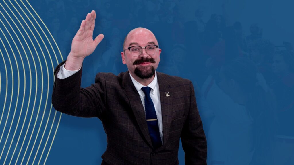 A thumbnail with a cheerful person signing "hello" in sign language". They are bald, with a stylized goatee, grey suit, white shirt, and blue tie. The background is a photo with blue overlay. The left side has swooping yellow lines.