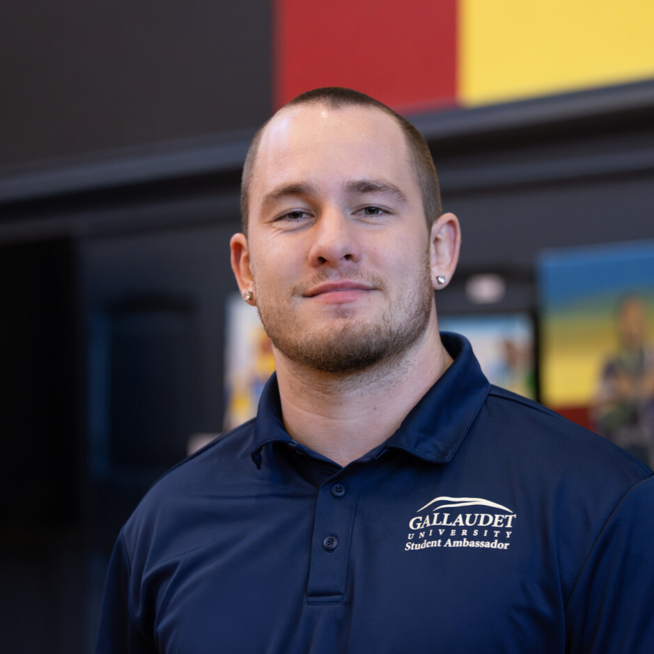 A photo of a college student smiling for the camera. They have shaved brown hair and facial hair. They wear a dark blue shirt with the Gallaudet University logo and text: "Student Ambassador".
