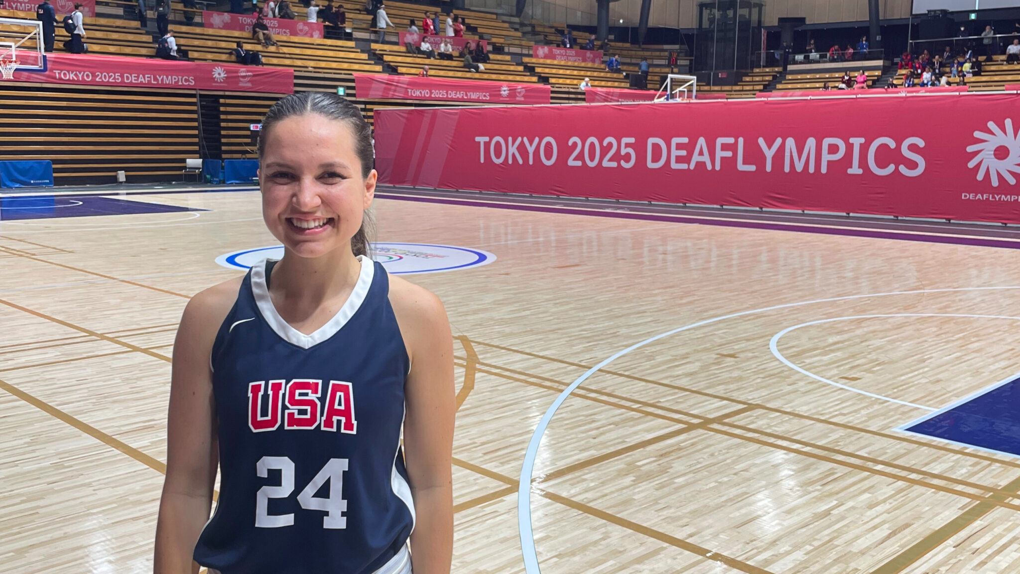 A women's basketball player stands on an empty court and smiles for the camera. They are wearing a dark blue jersey with "USA" and the number 24. On a partition wall behind the far sideline is text: "Tokyo 2025 Deaflympics".