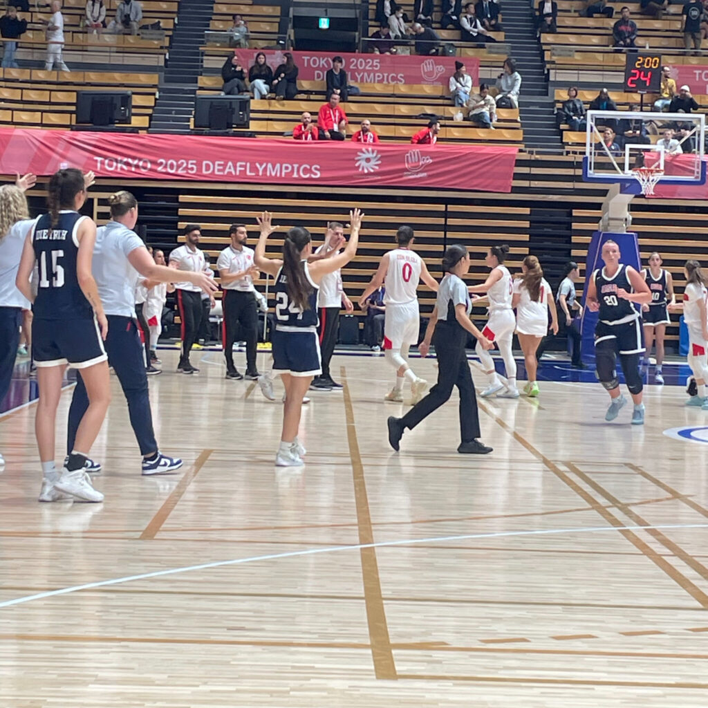 A photo of a women's basketball game while teams exit the court after a called timeout. In the center of the image, a player in a dark blue jersey walks from the sideline to their exiting teammates, offering a high five.