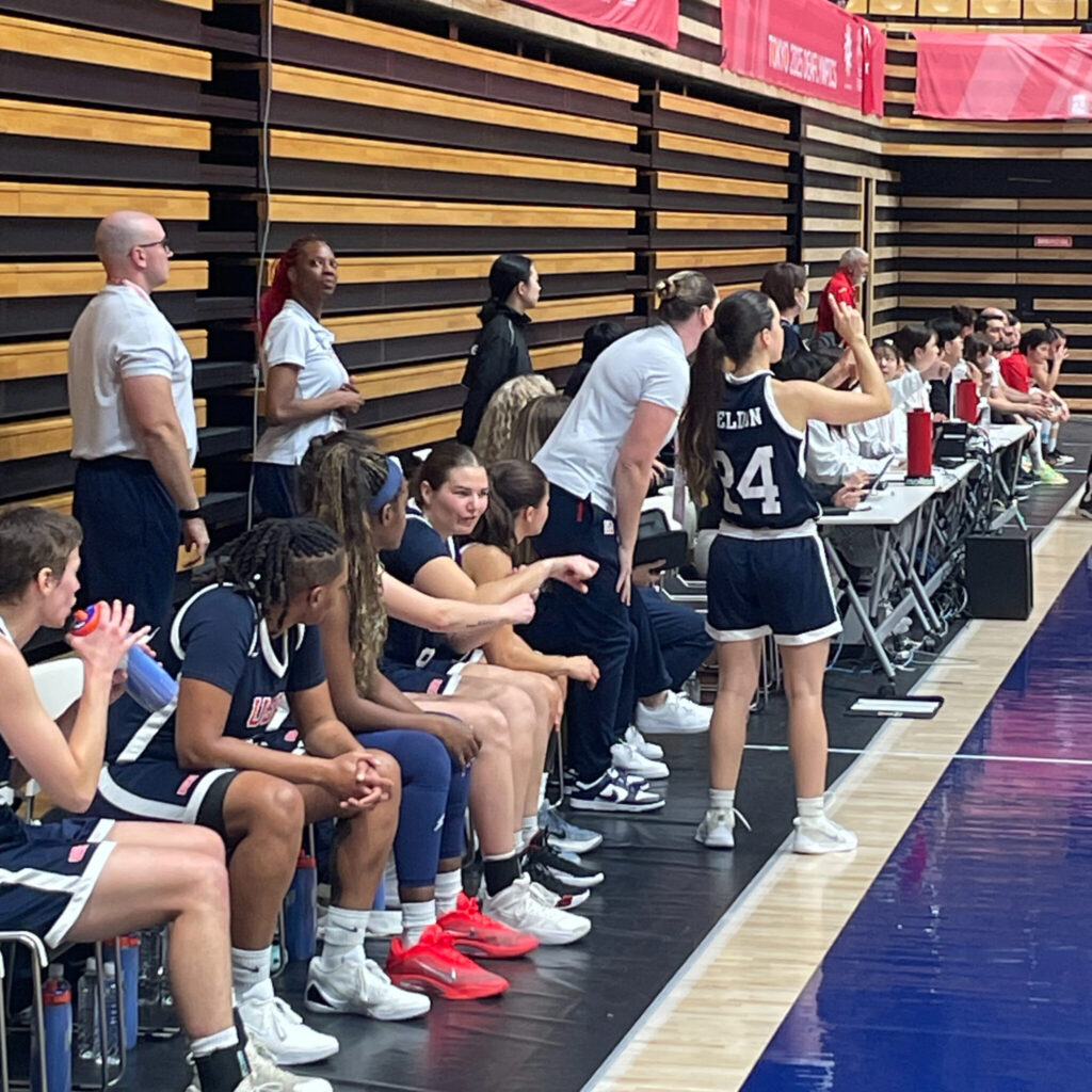 A photo of the sideline of during a women's basketball game. The team in view is wearing dark blue jerseys. One player is standing and communicating with teammates on the court, offscreen to the right. Their jersey has an obscured name and the number 24.