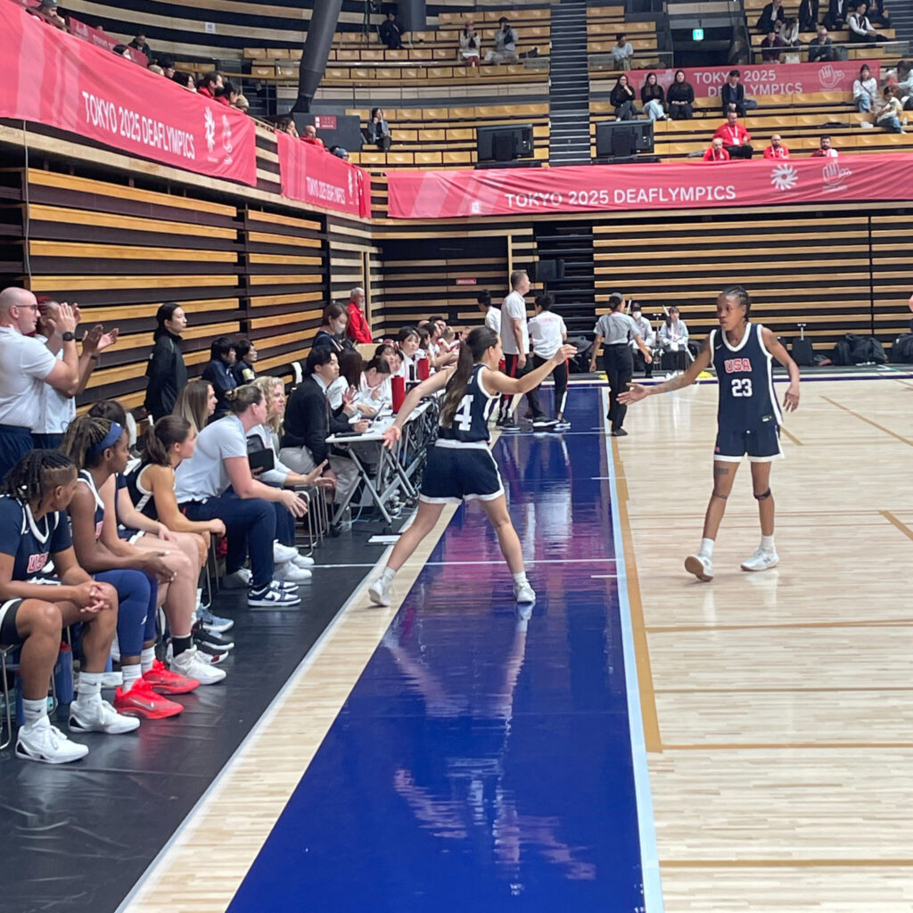 A photo of the sideline of during a women's basketball game. The team in view is wearing dark blue jerseys. In the center of the image, one player (number 24) is stepping off the bench to slap the hand of a teammate (number 23) walking off the court. A banner in the background says "Tokyo 2025 Deaflympics.
