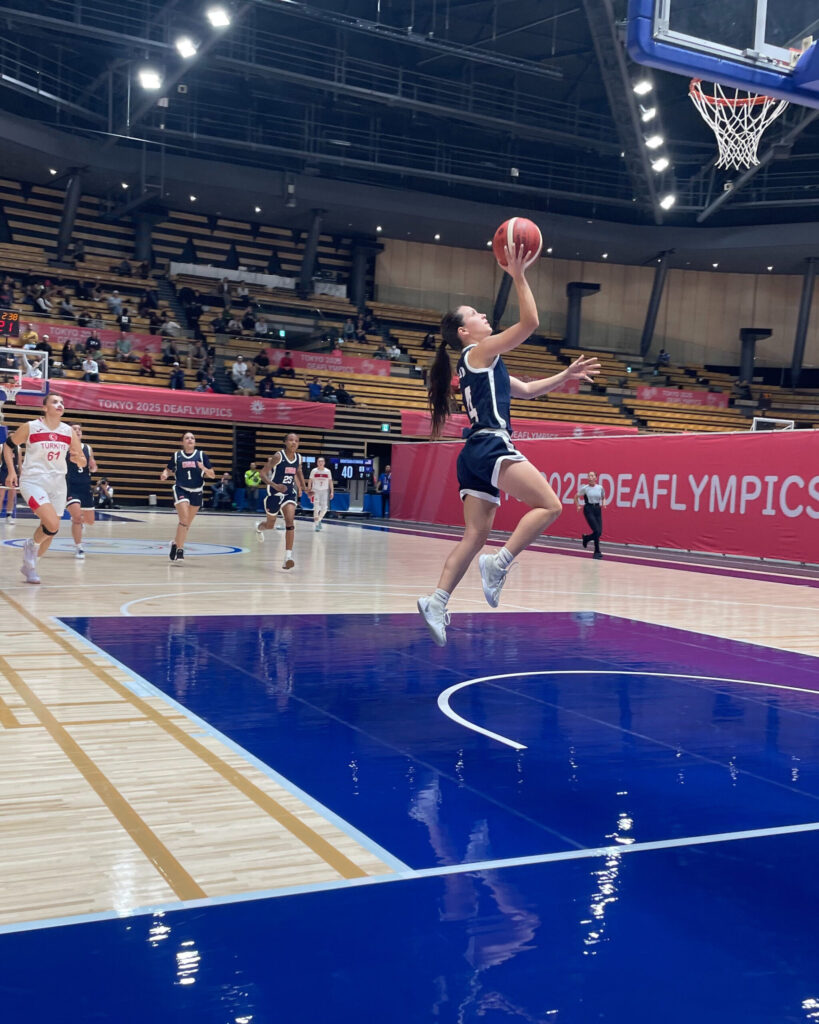 A women's basketball player jumps into a layup. They wear a dark blue jersey. Other players run up the court, watching the layup.