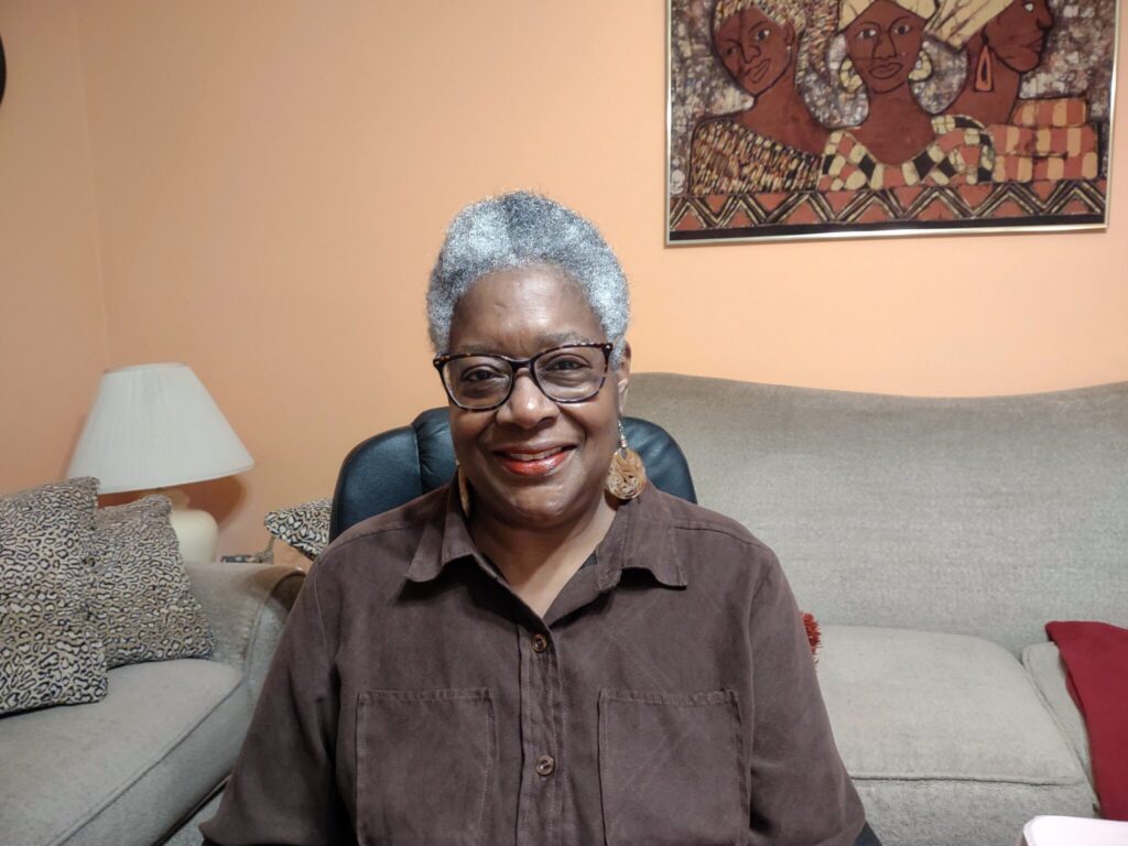 Image of woman with short gray hair and glasses seated in a room with gray couches and assorted throw pillows. She wears wooden earrings and a brown button down blouse.