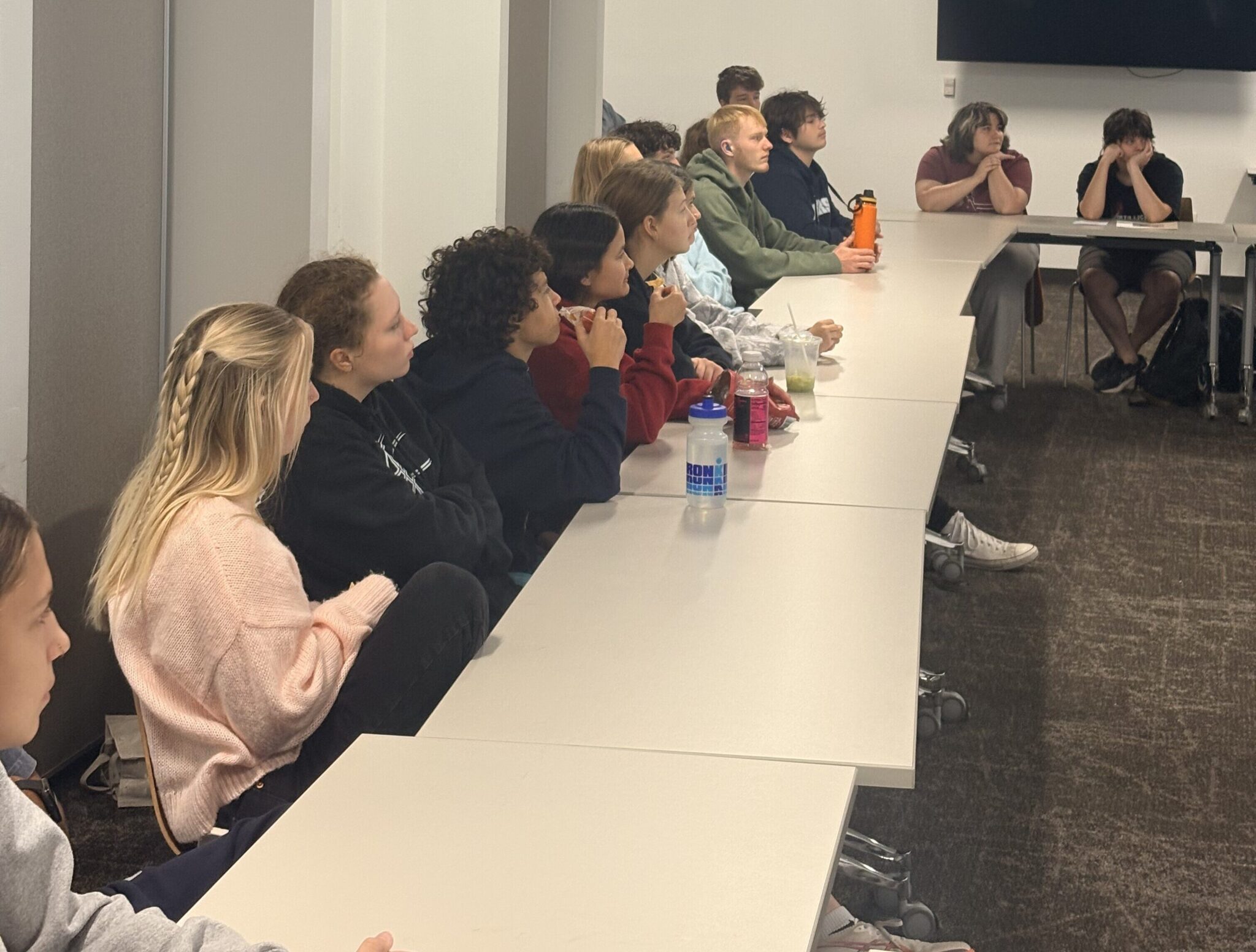 About a dozen teenagers sit at a long white table. They seem engaged by what they are watching in front of them.