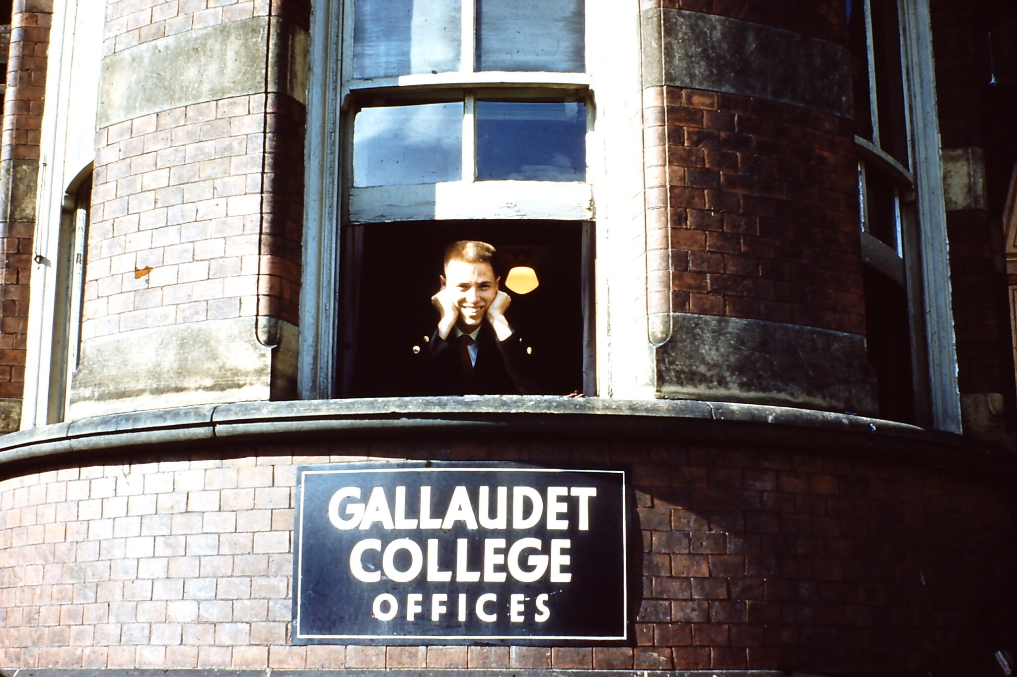Man sticks his head out of a window of a brick building. There is a sign just below that reads, "Gallaudet College Offices"
