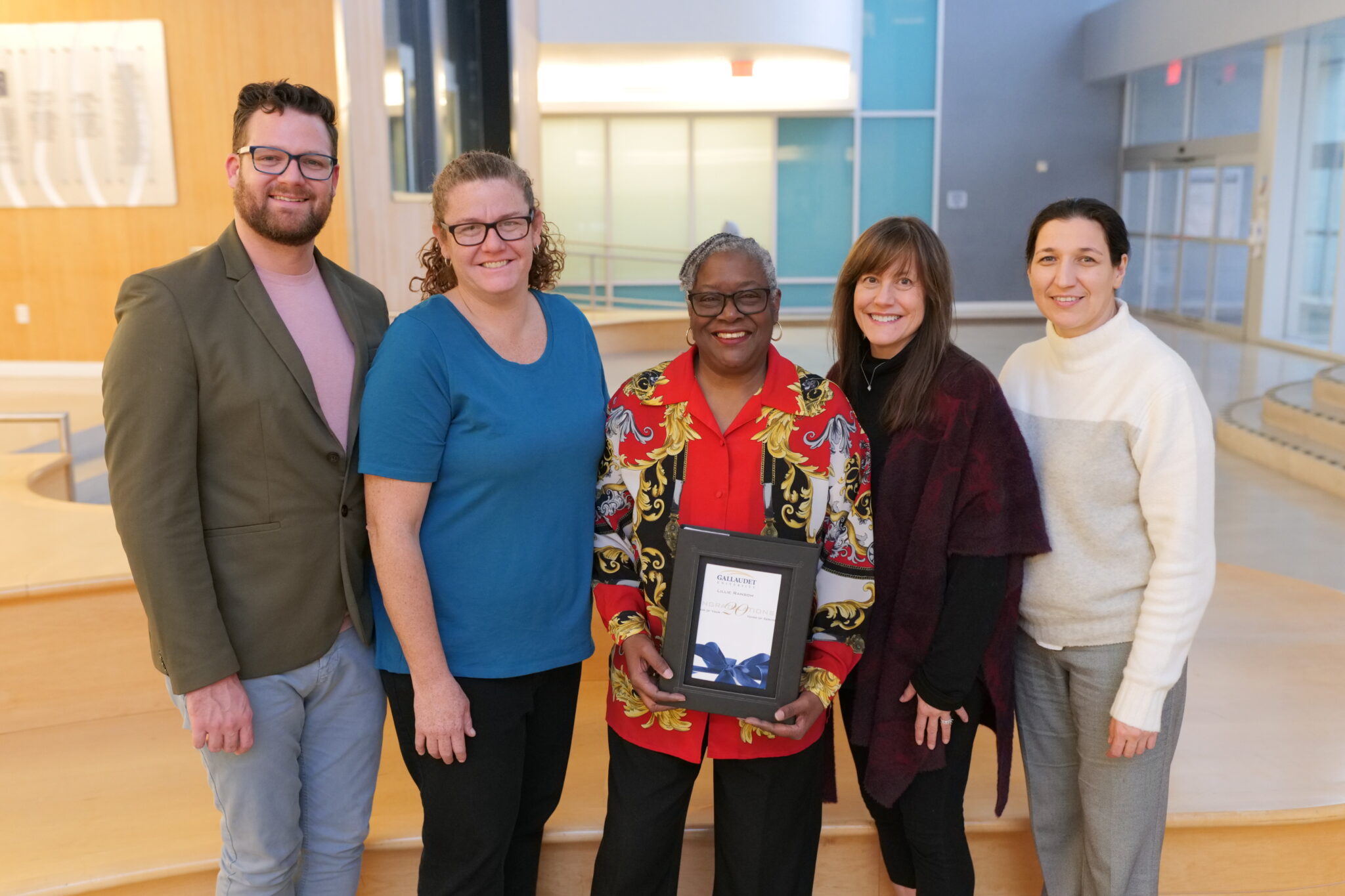 Five people stand in a row in a bright and open room. The woman in the middle is holding a black box.