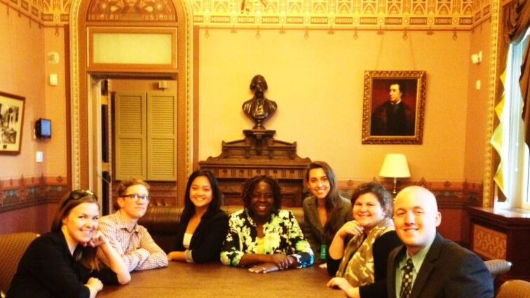 A photo of seven people sitting around the end of a large table, smiling for the camera. They are in a stately room. In the middle of the image, directly behind the group, is a desk with a sculpted bust on top. To the right, on the wall, is an old painted portrait.