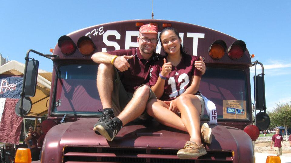 A photo of two people on the hood of a maroon bus, smiling and giving a thumbs up for the camera. The person on the left has their left arm around the person on the right. They both wear maroon. The brim of the bus has obscured text: "The Spirit".