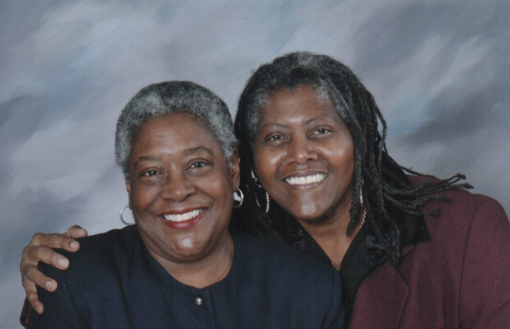 Two smiling women sit next to each other. The woman on the right has her arm around the woman on the left.