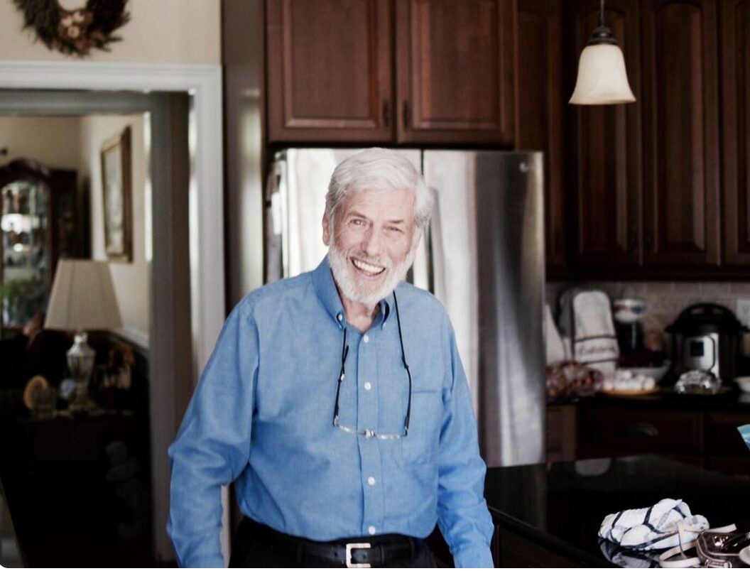 A man with gray hair and facial hair stands in a kitchen in front of a refrigerator. He wears a button down blue shirt and has a pair of glasses hanging around his neck