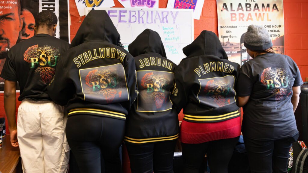 Five people stand side by side, facing away from the camera, each wearing black hooded sweatshirts with colorful designs on the back. The designs include the letters "PSU" and names that appear to be individual identifiers. The background features a bulletin board with posters and decorations related to an event or celebration.