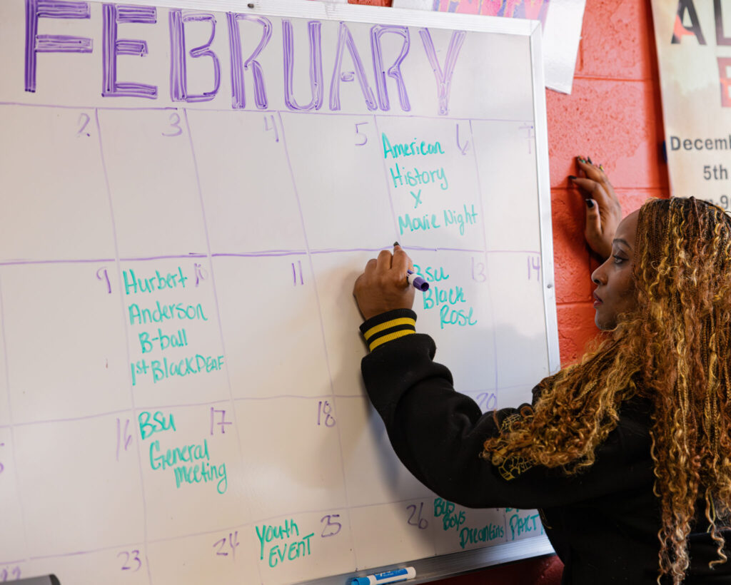 A person with long, curly hair is writing on a whiteboard calendar for February. The calendar has dates and events listed, including "American History" and "Youth Event." The background features a red wall.