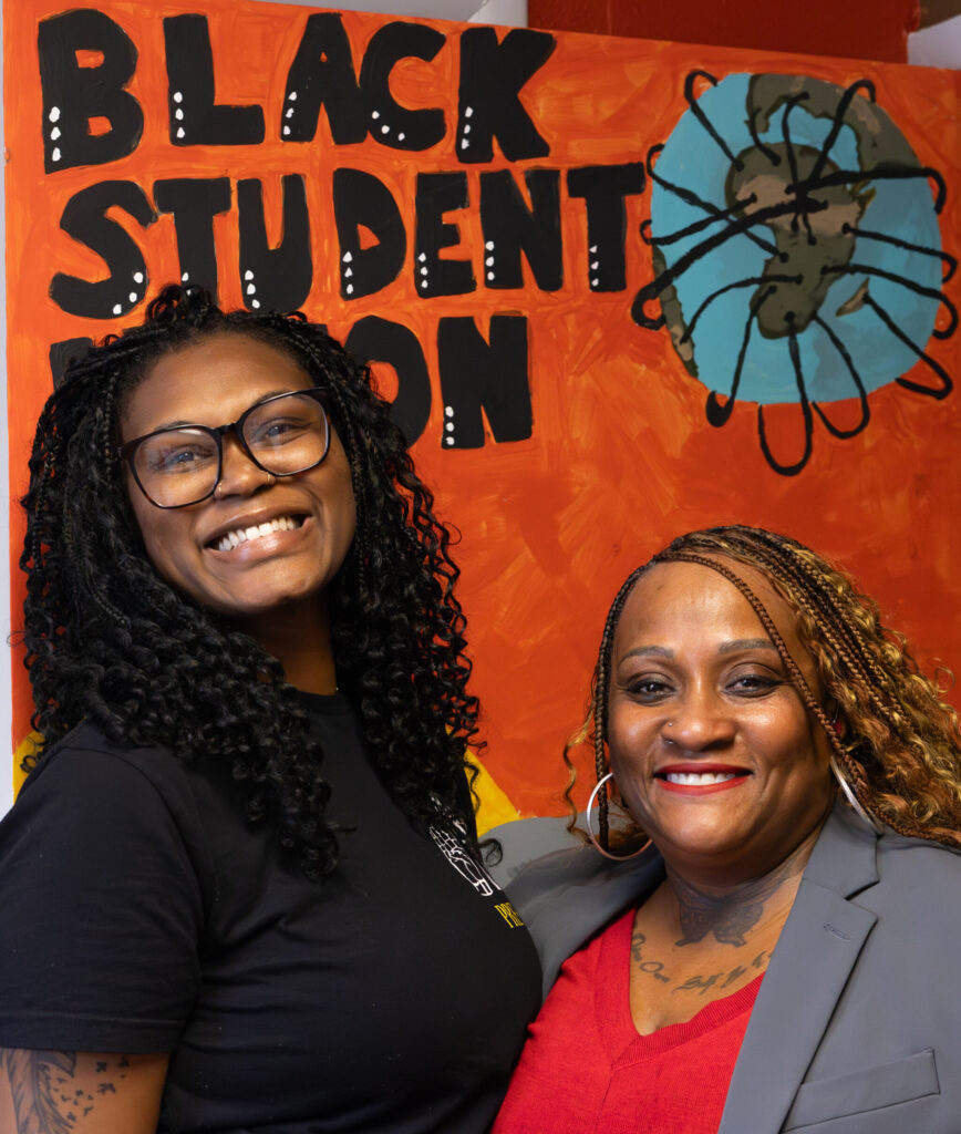 A smiling woman with curly hair and glasses stands alongside another woman with long, styled hair, both posing in front of a colorful backdrop that reads "BLACK STUDENT UNION." The backdrop features a graphic of a globe.