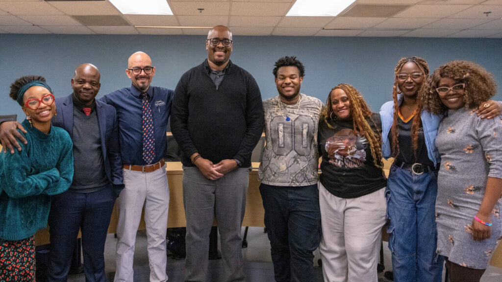 A photo of eight people in a university room smiling for the camera. They are dressed in a variety of different clothing, from casual to formal. The person fourth from left is notably tall, while the others are various heights.