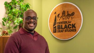 A photo of a person smiling for the camera in an office. They are bald, with glasses and a red sweater. On the right side of the image, slightly out of focus in the background, is a circular sign with the logo for the Center of Black Deaf Studies.