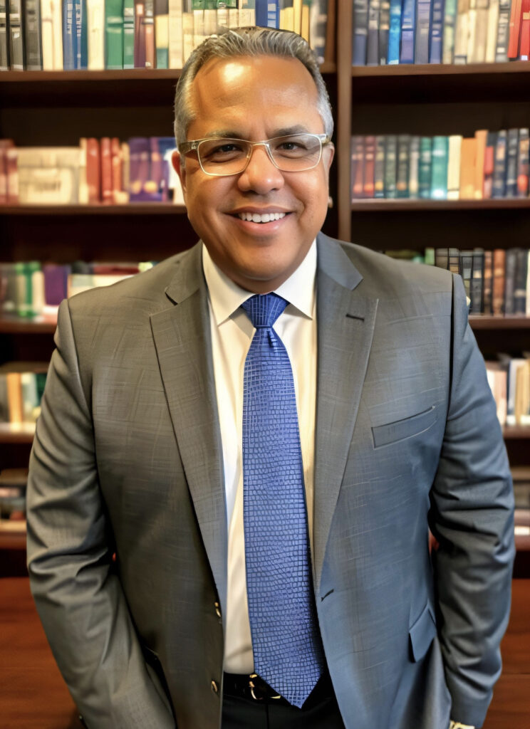 A man in glasses and a gray suit stands in front of a bookcase.