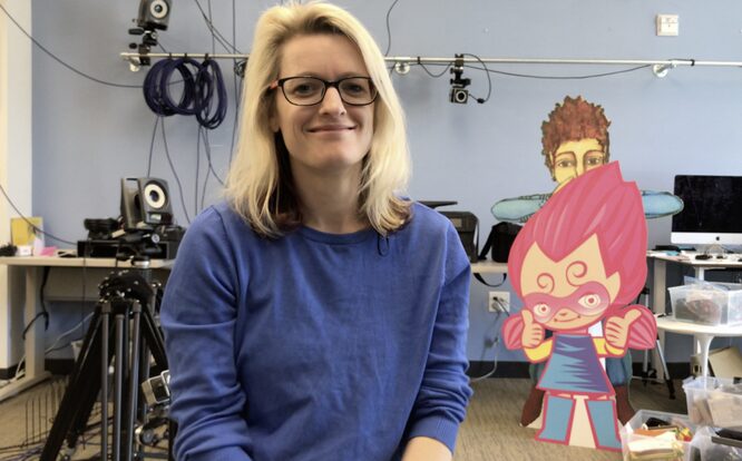 Woman with blond hair and glasses sits in a room with filming equipment. There's a cardboard cutout next to her of a girl in a blue dress with pink hair.