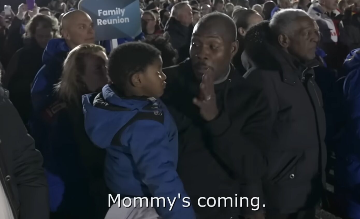 Image of a man holding a young boy in the middle of a large crowd at night. The man is bringing one hand to his chin. Underneath, text reads, "Mommy's coming."