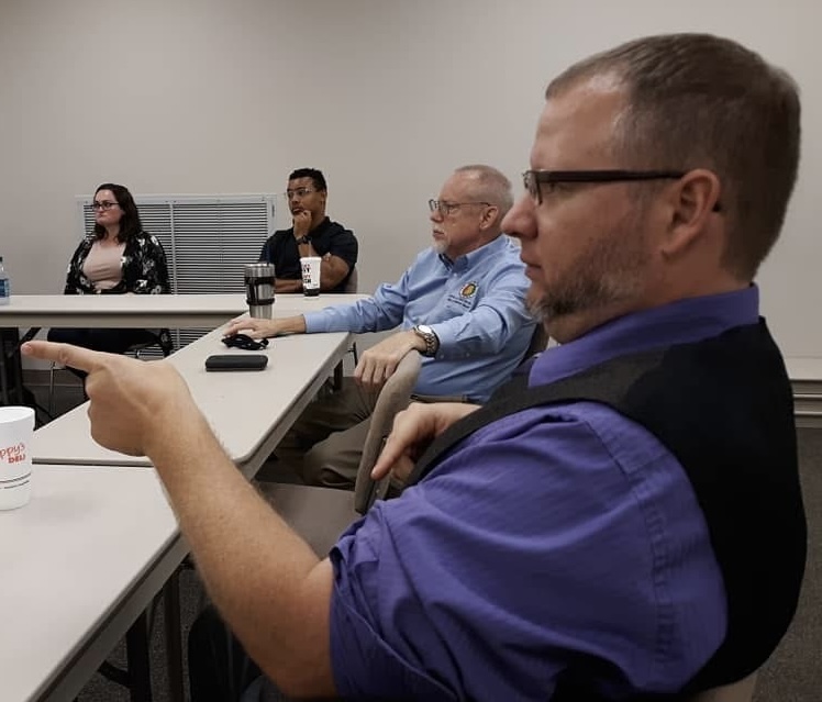 Man in a purple shirt and dark vest is seated at a long white table and seen in profile pointing in front of him. There are three other people sitting at the table in the background. All are looking at something off to the left.