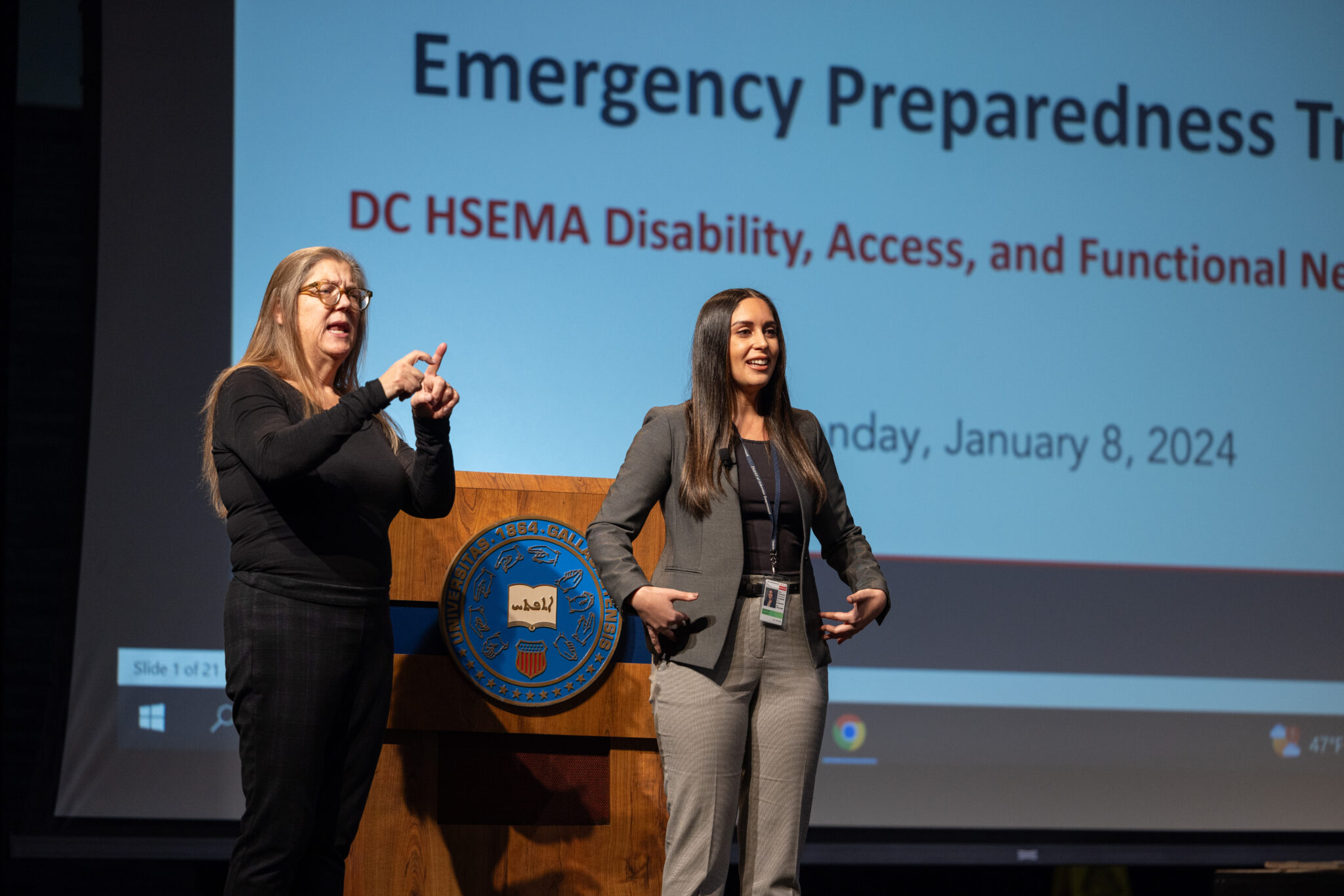 Two women are standing on a stage during a presentation titled "Emergency Preparedness Training." One woman is using sign language, while the other is speaking. A large screen in the background displays the title and date of the event, which is focused on disability, access, and functional needs in emergency management. A podium with a logo is visible in the foreground.