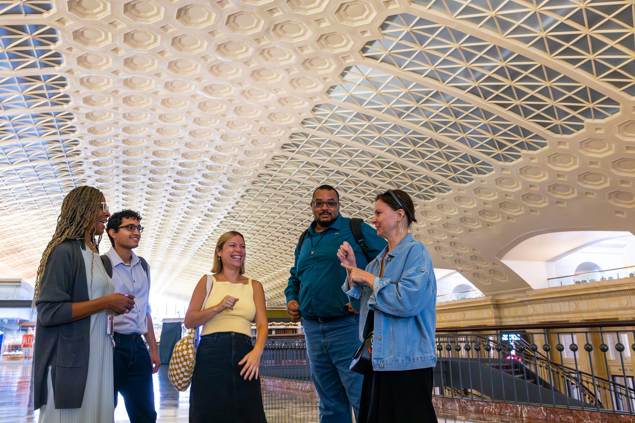 A group of five people stands together in an indoor space with a decorative ceiling. They appear to be engaged in conversation, with one person gesturing while speaking. The atmosphere is bright and lively, highlighting the architecture of the space.