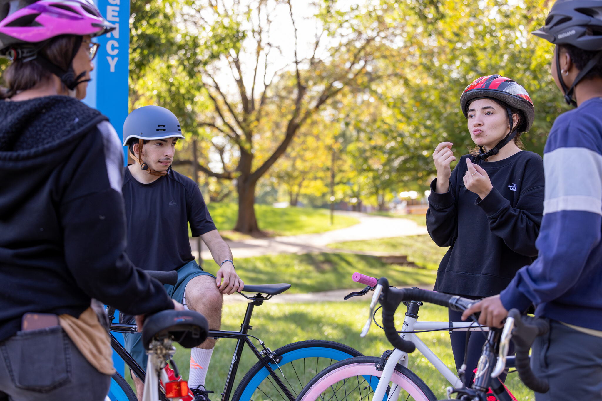 A group of four cyclists wearing helmets is gathered in a park, engaging in conversation while standing next to their bicycles. The setting features lush greenery and trees in the background, indicating a sunny day. One person is gesturing while another appears to be listening, and the group seems to be enjoying their time outdoors.