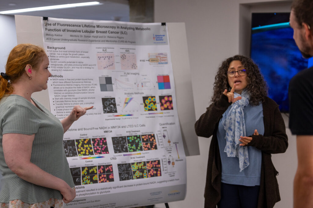 Two women are discussing a research poster in a conference setting. One woman, with curly hair and glasses, is gesturing towards the poster while explaining. The other woman, with red hair, is listening attentively. The poster features colorful graphs and images related to scientific research. A man is partially visible on the right, observing the discussion.