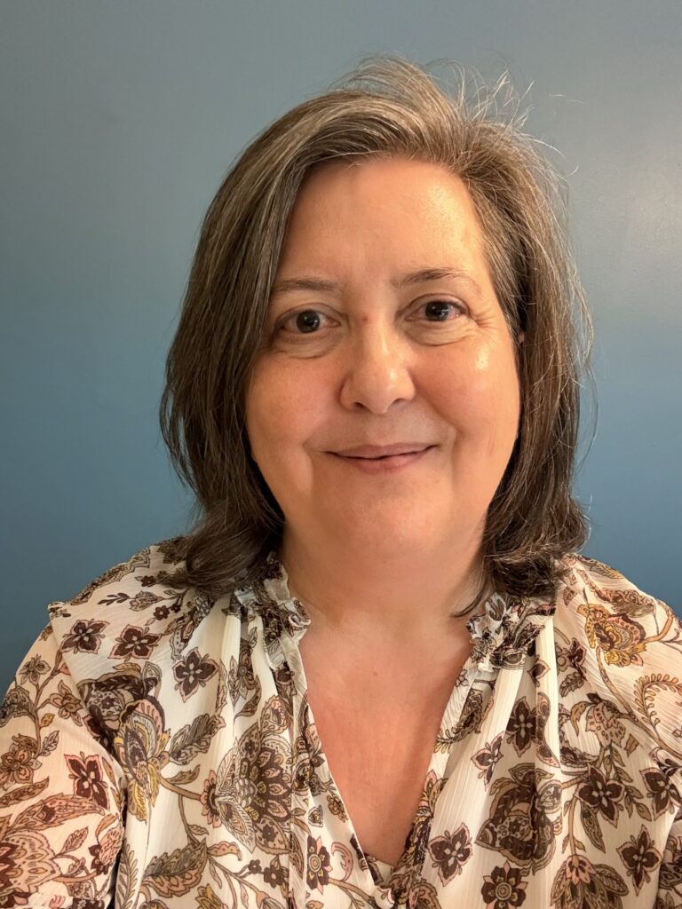 A woman with shoulder-length gray hair smiles at the camera, wearing a floral-patterned blouse. The background is a solid light blue.