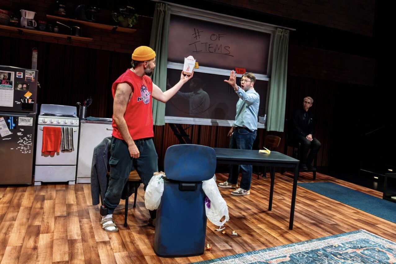 Two men stand in a stage set designed to look like a kitchen. A man in a red shirt and yellow hat holds up a Chinese takeout container toward another man in a blue dress shirt.