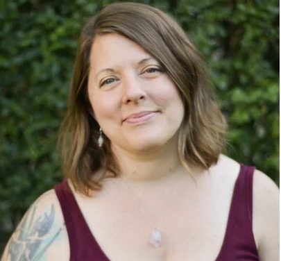 Image of a woman wearing a maroon tank top. She is posing in front of a large plant.