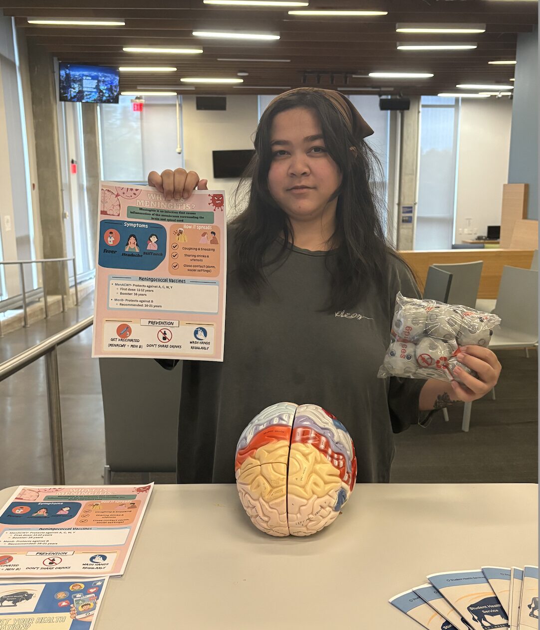 Woman inside an office has a model of the human brain in front of her. In one hand she holds up a sheet of paper with medical information. In the other hand, she has a bag.