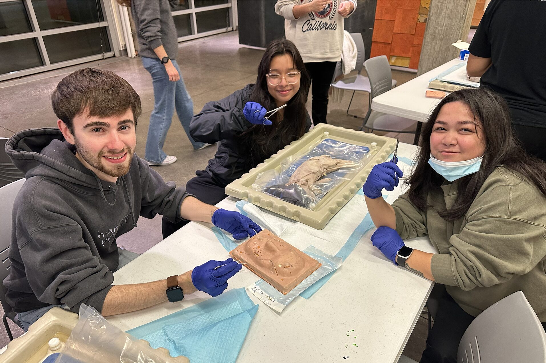 A young man and two young women are at a white table. There are some pink slabs in front of them. They are wearing medical gloves and holding medical instruments.