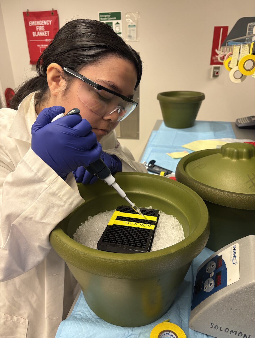 Woman in a lab coat and protective eye wear is wearing blue gloves and holding a stick-shaped object over a green bowl.