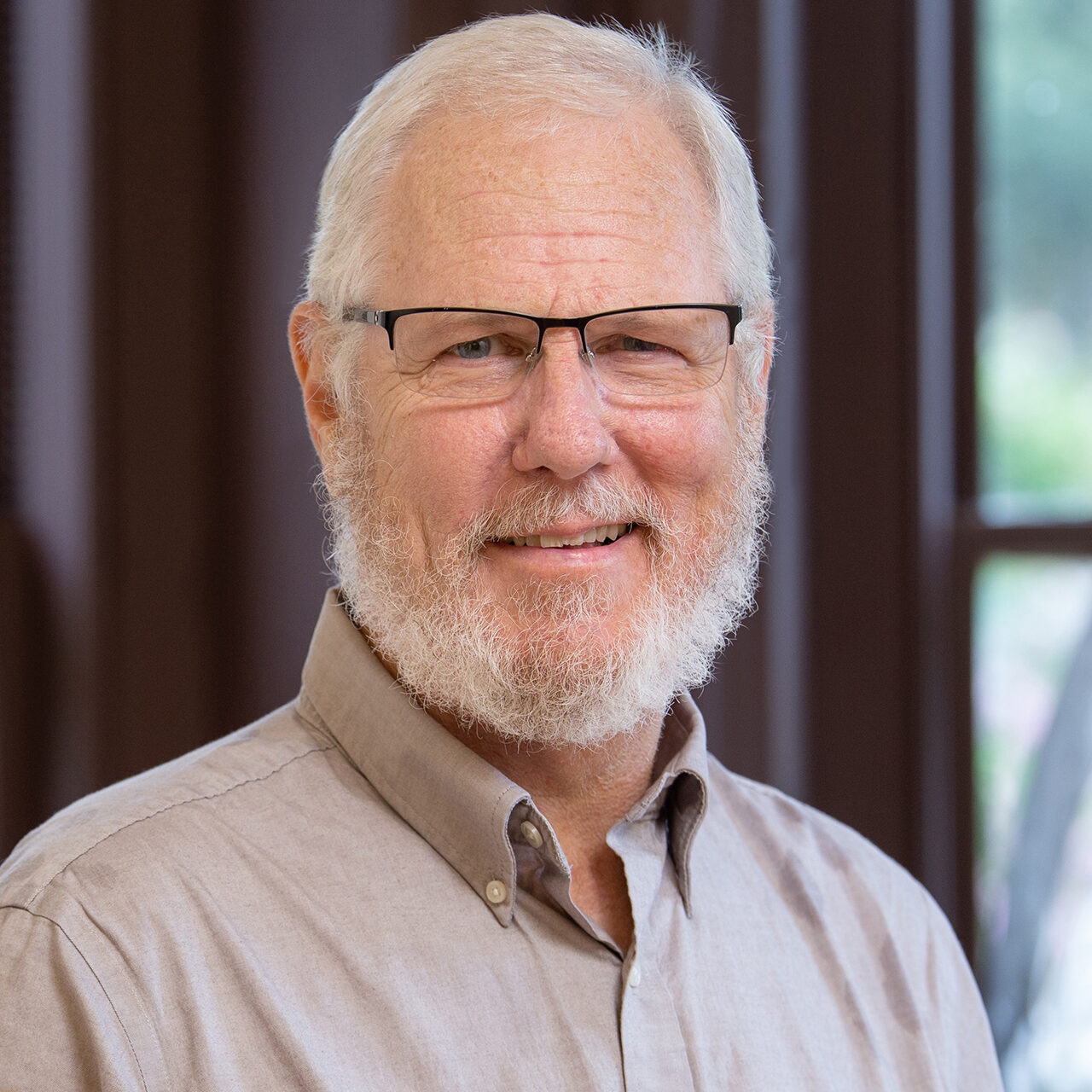 A smiling older man with a beard and glasses, wearing a light-colored button-up shirt, stands in front of a window with natural light.