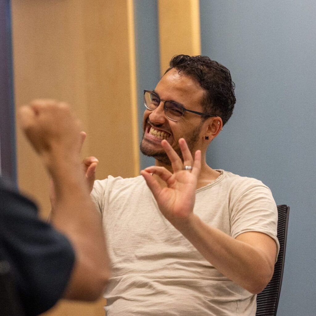 A man with glasses and a light-colored shirt smiles while gesturing with his hands, engaged in a conversation with another person who is partially visible in the foreground. The setting appears to be an office or meeting room with muted colors.