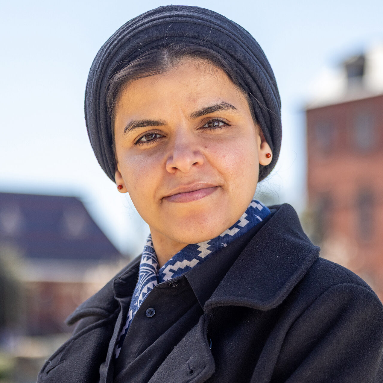 A woman with dark hair, wearing a black coat and a patterned scarf, stands outdoors with a slight smile, against a blurred background of buildings.