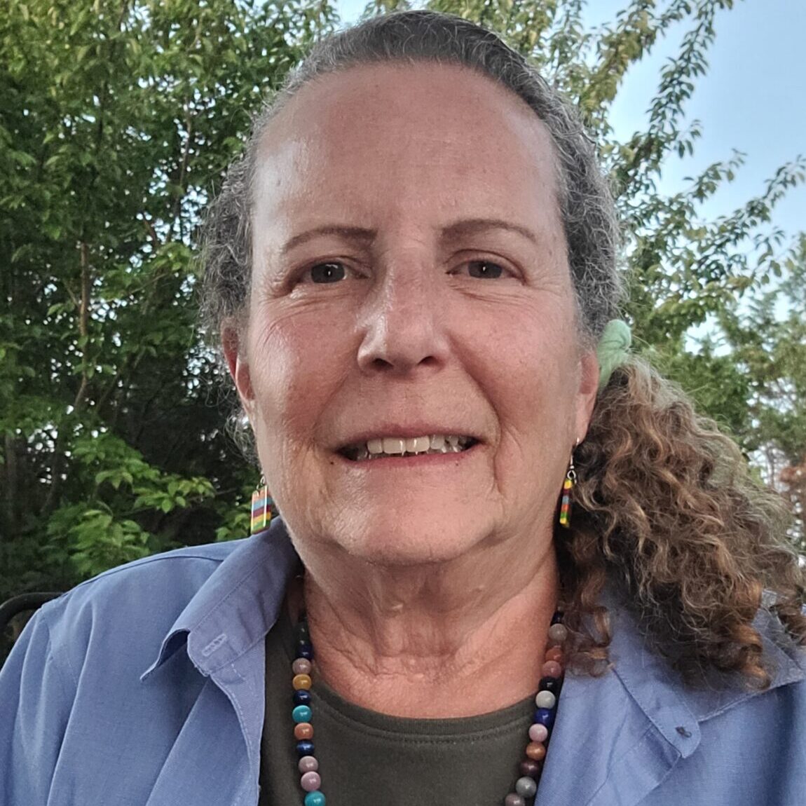 A smiling woman with long, curly hair, wearing a blue shirt and a beaded necklace, stands outdoors with greenery in the background.