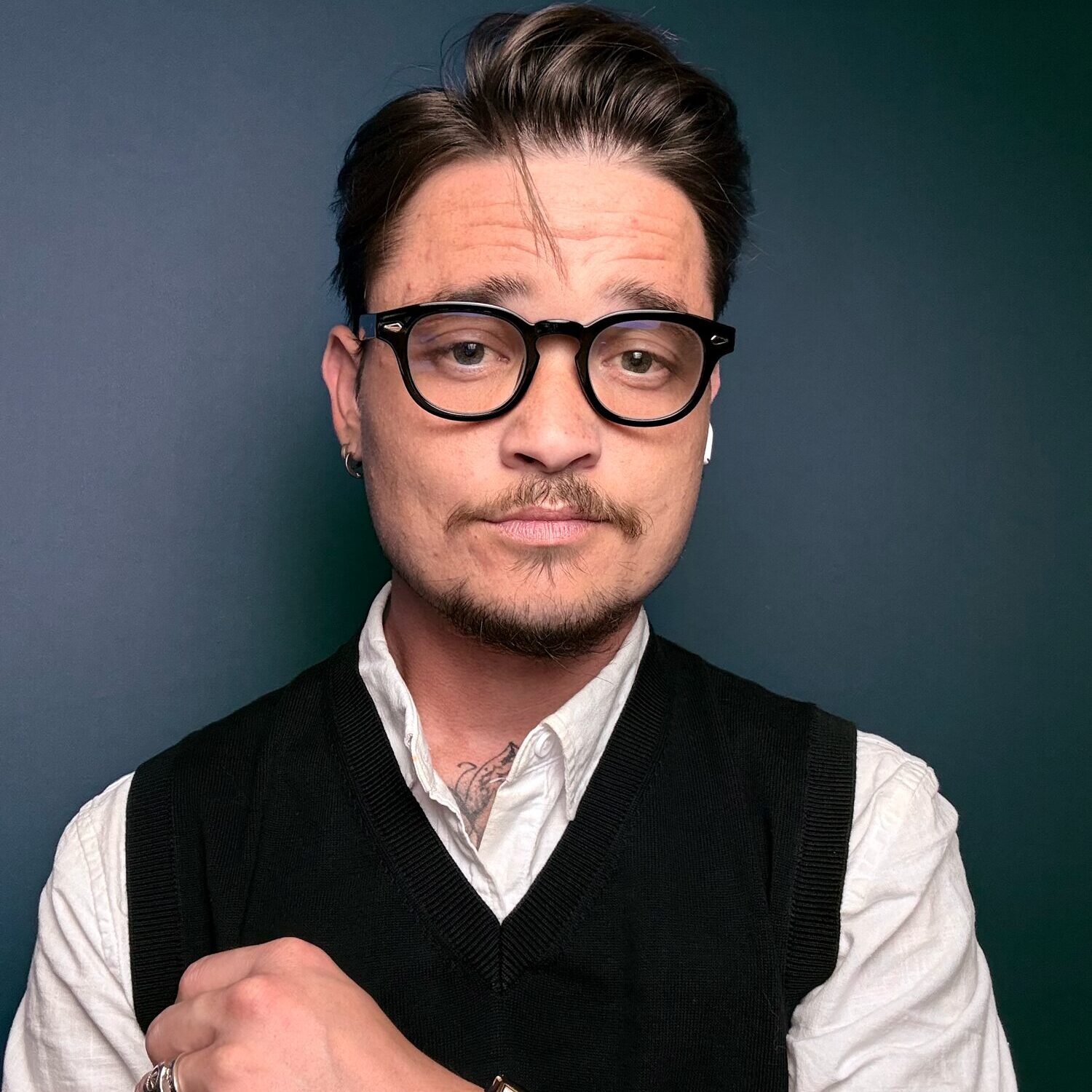 A person with short, styled hair and glasses is wearing a white shirt and a black vest. They are looking directly at the camera with a slight smile, showing confidence. The background is a gradient of dark green.