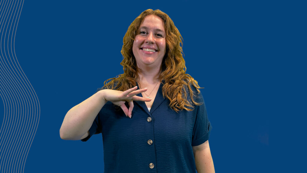 A person with long, wavy hair is smiling and making a hand sign while wearing a dark blue shirt, set against a solid blue background.