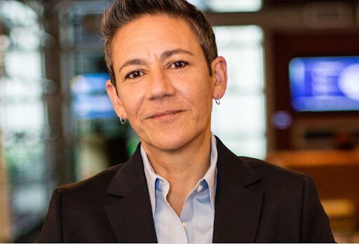 Dr. Maribel Gárate-Estes is standing against a blurred background. Dr. Gárate-Estes has short dark hair and is wearing a navy blue jacket over a light blue button down shirt