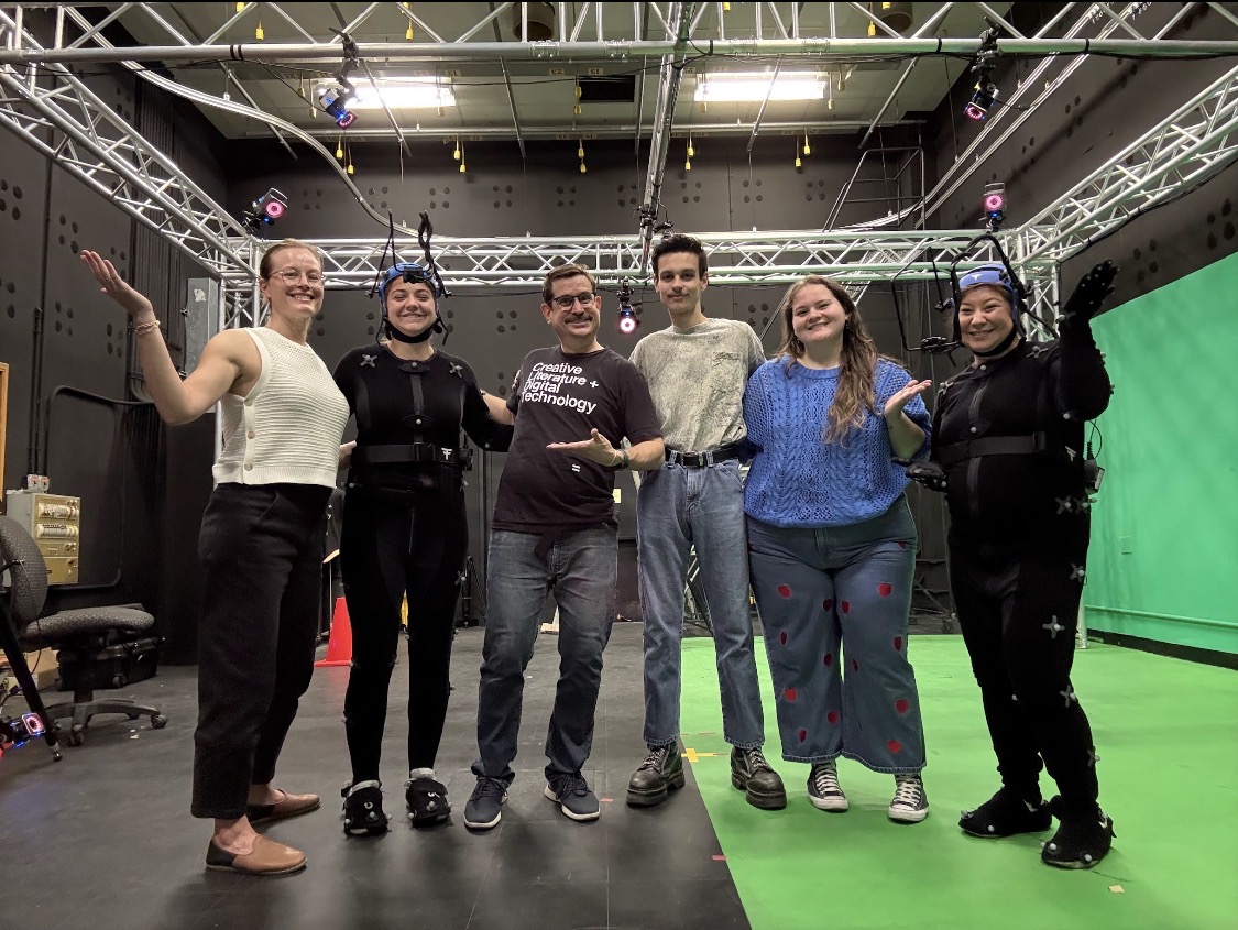 A group of six people stand in a black, industrial looking room. Half of the floor is green and there is a green screen off to the right side.