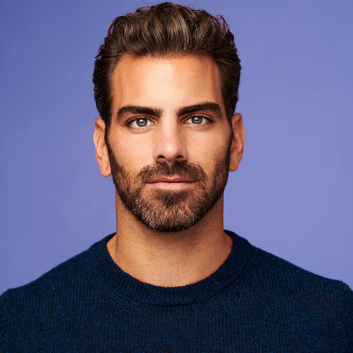 A close-up portrait of a man with short, styled hair and a well-groomed beard, wearing a dark blue sweater. He has a serious expression and is facing the camera against a soft purple background.