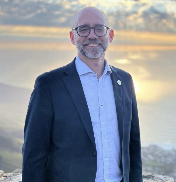 Light breaks through clouds in the background of a man in light blue shirt and navy blazer