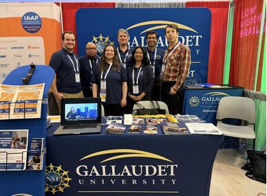 Seven people stand behind a table draped in a navy blue cloth that has "Gallaudet" on it. There are papers on the tables. Behind them is a banner that also reads, "Gallaudet"