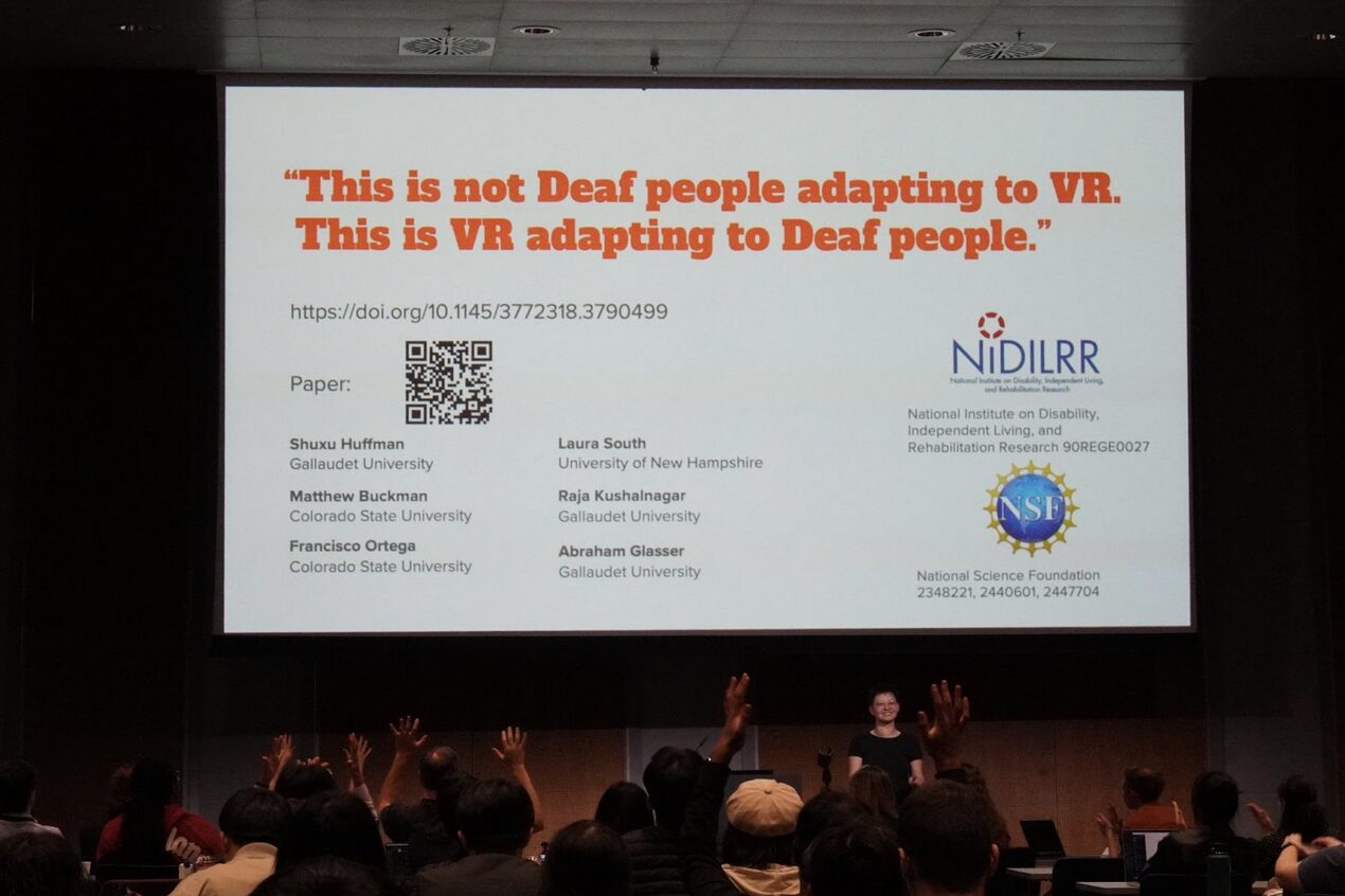 A large screen has bold orange text that reads, "This is not Deaf people adapting to VR. This is VR adapting to Deaf people." There is other text below. The backs of the heads of people in the audience are in the foreground.