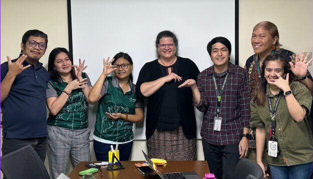 A group of six people standing in front of a whiteboard, smiling and making various hand gestures. They are dressed casually, with one person holding a notebook. The setting appears to be an office or meeting room.