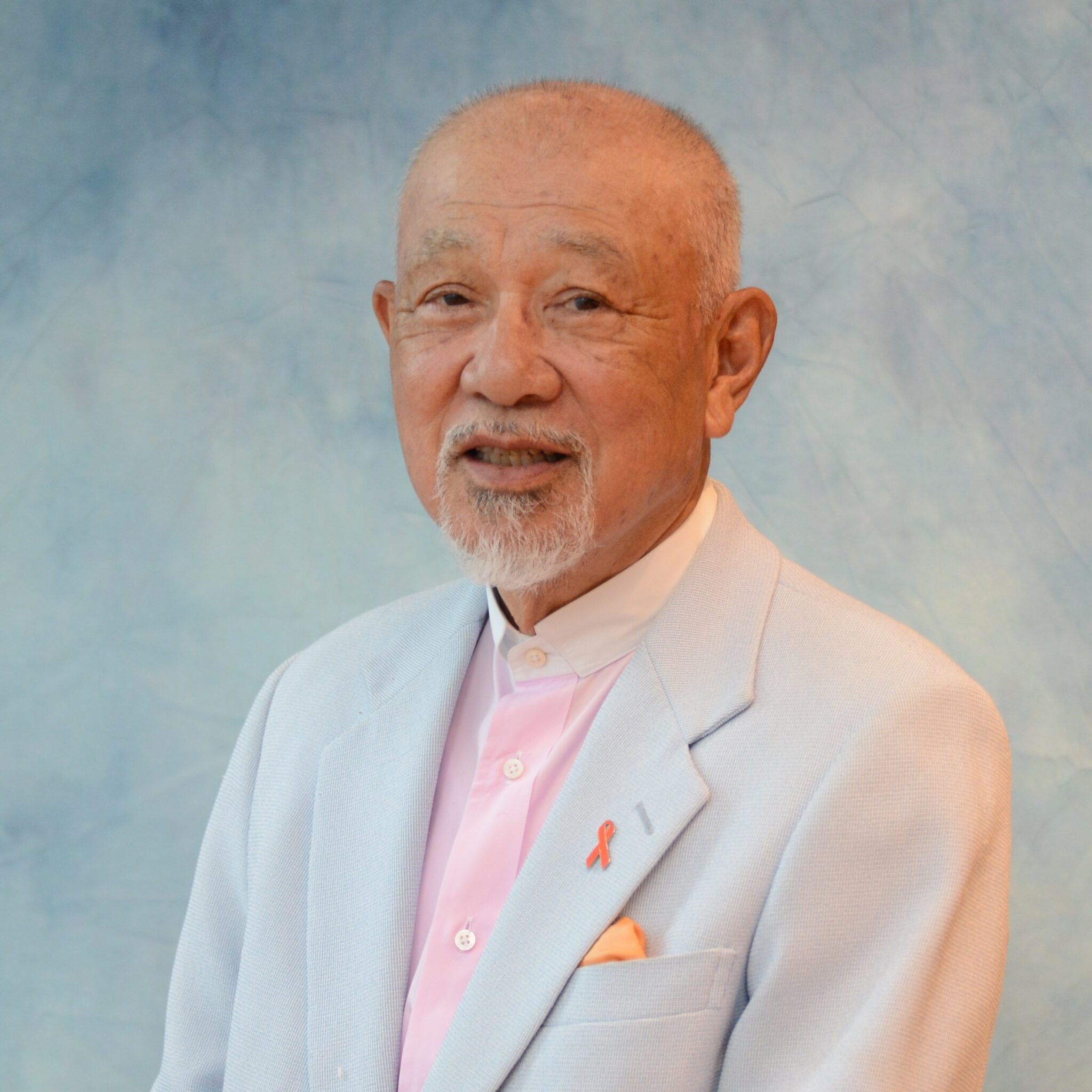 A smiling elderly man with a short beard, wearing a light blue suit jacket over a pink shirt. He has a small orange ribbon pinned to his lapel and is set against a soft blue background.