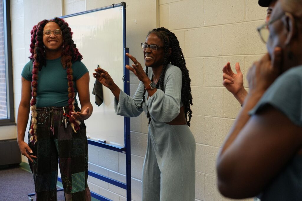 Two women are engaged in a lively conversation in a classroom setting. One woman, with long curly hair and wearing a teal top and patterned pants, smiles as she listens. The other woman, with long straight hair and wearing a light blue outfit, gestures animatedly while speaking. A whiteboard is visible in the background, and a couple of other individuals can be seen partially in the frame, contributing to the dynamic atmosphere.